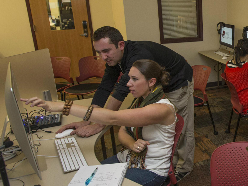 A student is seated at her computer, and her professor points to her screen